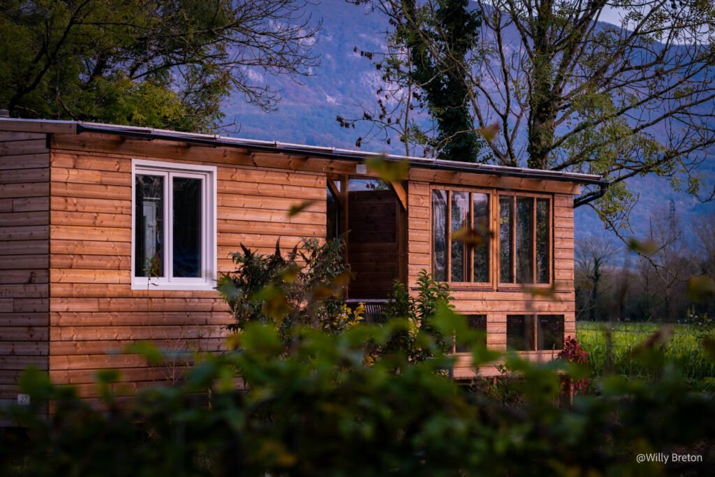 Maison en bois écologique au coucher du soleil, entourée de verdure et située dans un cadre naturel paisible avec des montagnes en arrière-plan