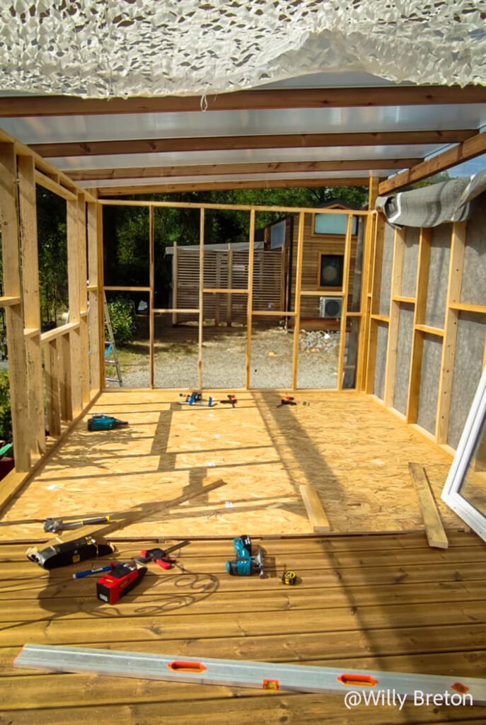 Travaux de construction en bois, avec outils et équipements sur une terrasse en bois, sous une couverture semi-transparente.