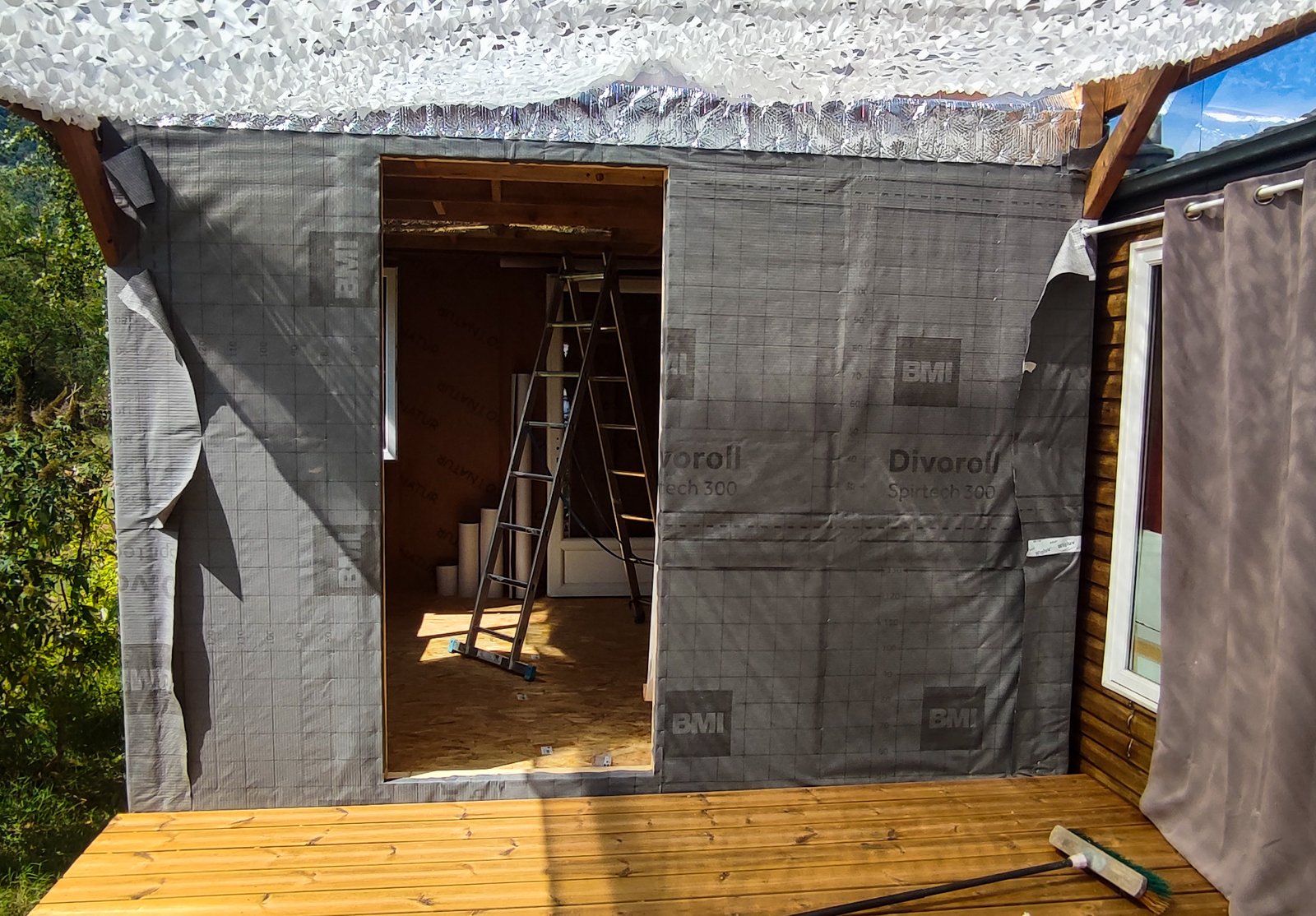 Construction en cours avec une ossature en bois, revêtement installé et terrasse en bois, vue sur l'intérieur à travers une ouverture.
