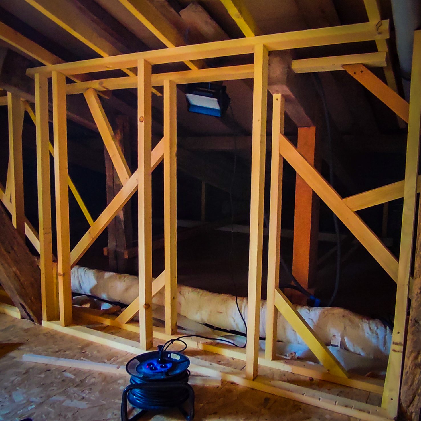 Charpente en bois pour cloison dans des combles en rénovation, avec lumière de chantier et câblage en cours.