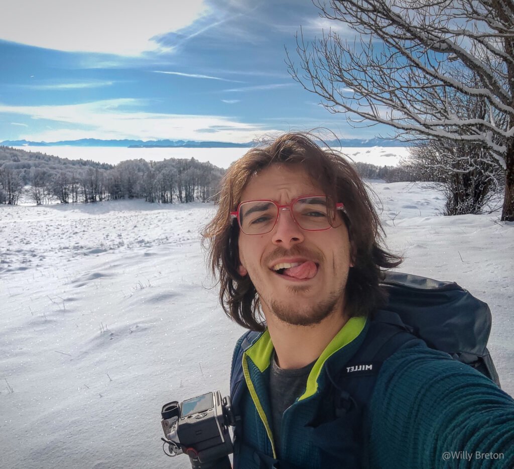 Portrait d'un randonneur(Willy Breton) souriant avec sac à dos, prenant un selfie au milieu d'un paysage enneigé, sous un ciel bleu dégagé.