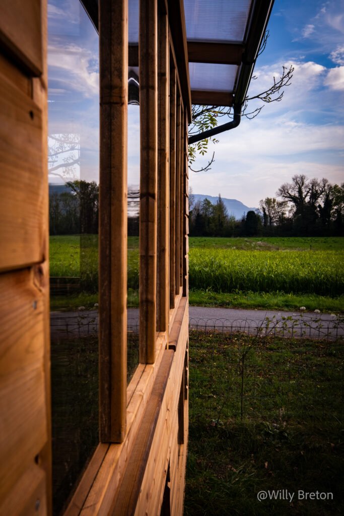 Façade en bois avec grandes ouvertures vitrées, offrant une vue sur un paysage naturel de champs et montagnes sous un ciel dégagé.