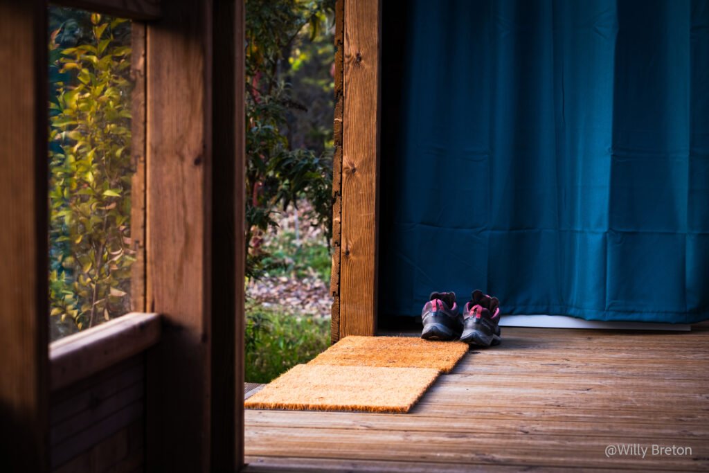 Paire de chaussures posées sur un tapis devant une entrée en bois, donnant sur un espace naturel, symbole de simplicité et de connexion avec la nature.