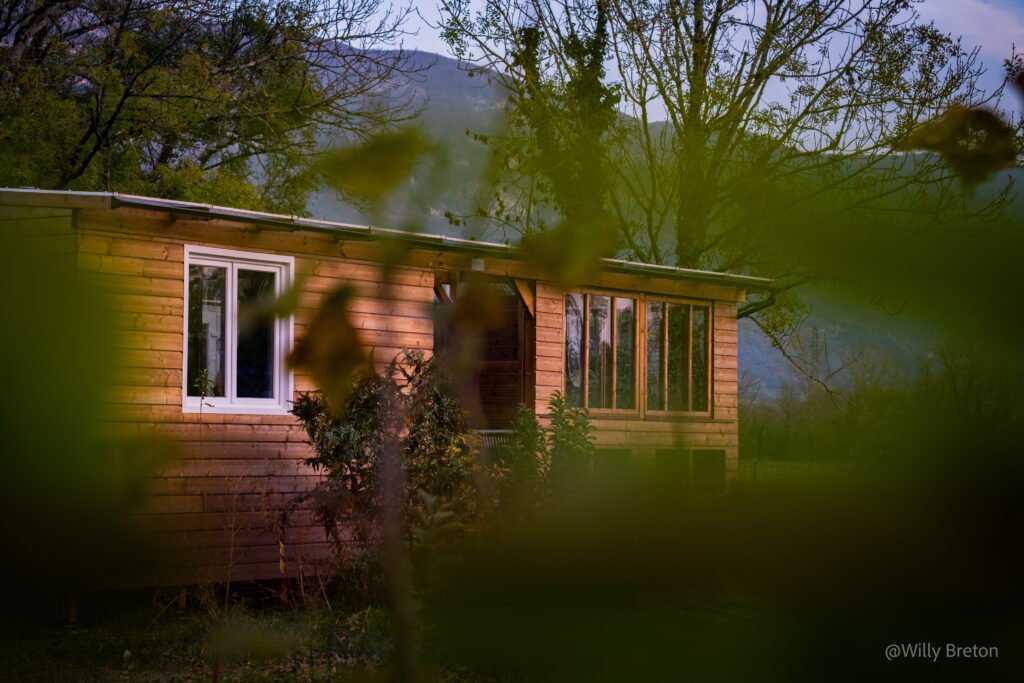 Vue d'une maison en bois entourée de végétation, intégrée dans un cadre naturel avec des montagnes en arrière-plan.