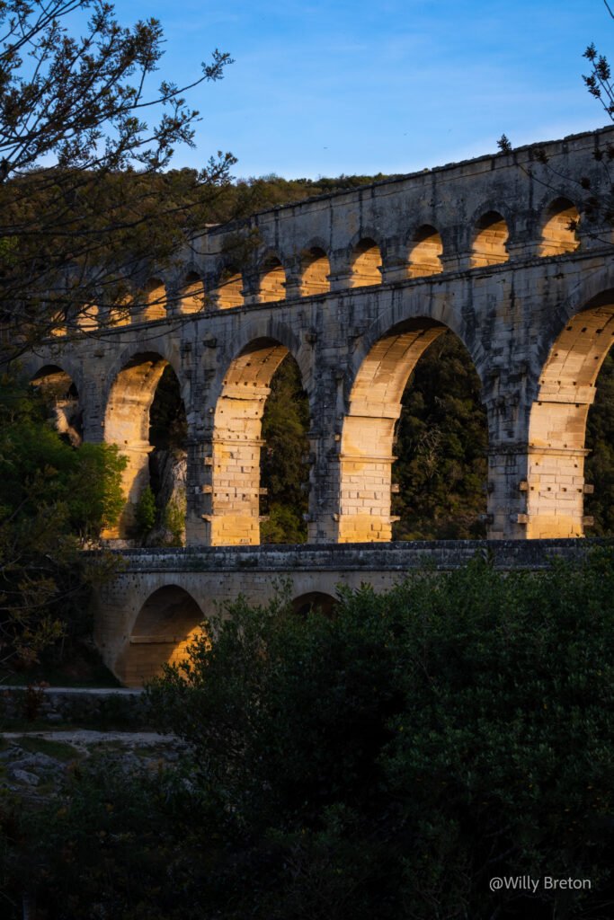 Vue du Pont du Gard au coucher du soleil, illuminé par une lumière dorée, entouré d’arbres et de végétation.