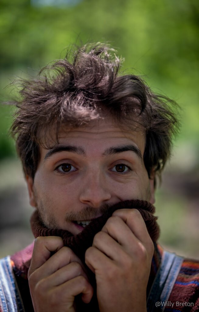 Portrait d’un homme avec un sourire naturel, enveloppé dans un pull, avec un arrière-plan flou de verdure.