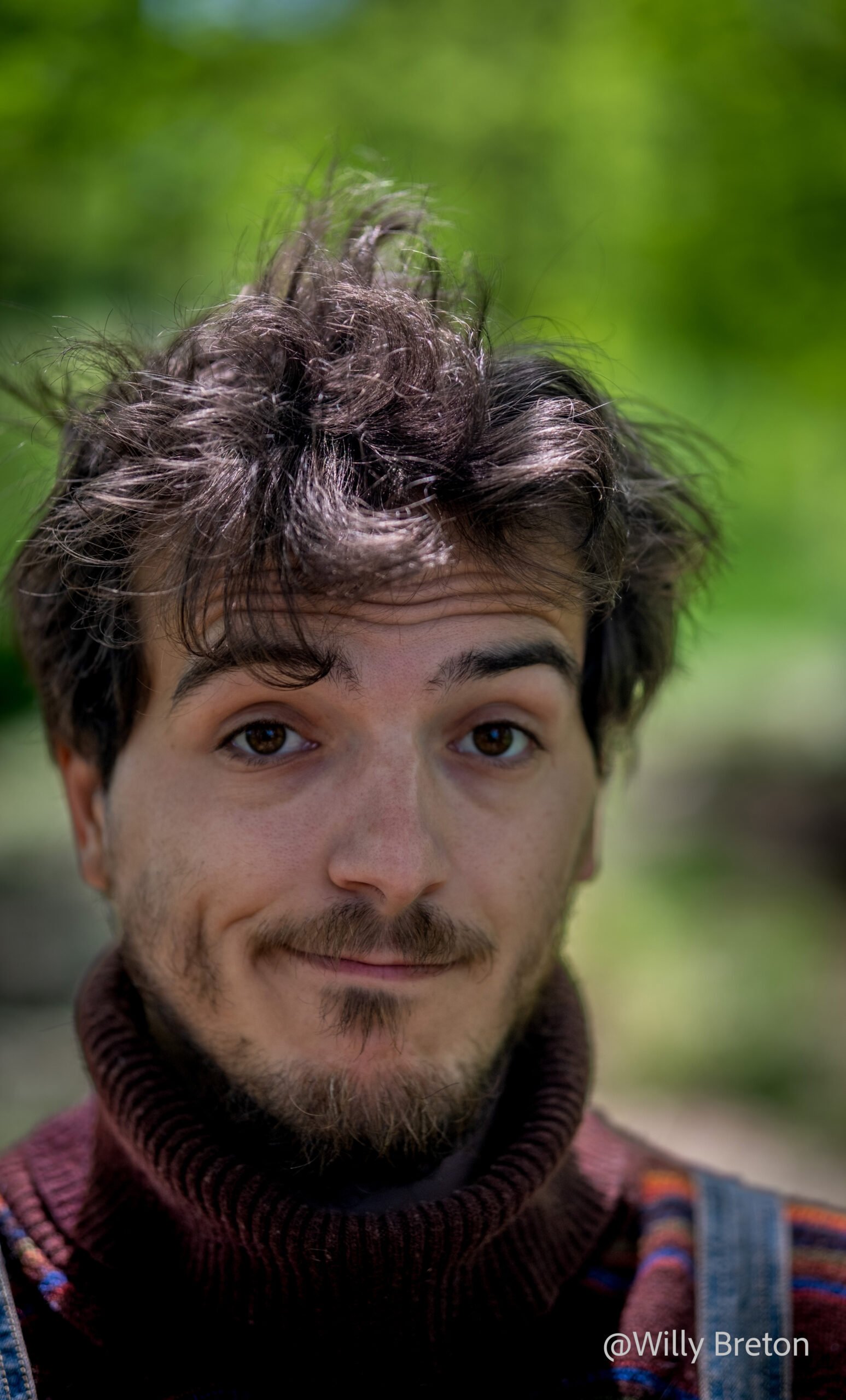 Autoportrait d’un homme avec un léger sourire et un regard expressif, capturé en plein air avec un arrière-plan naturel flou.